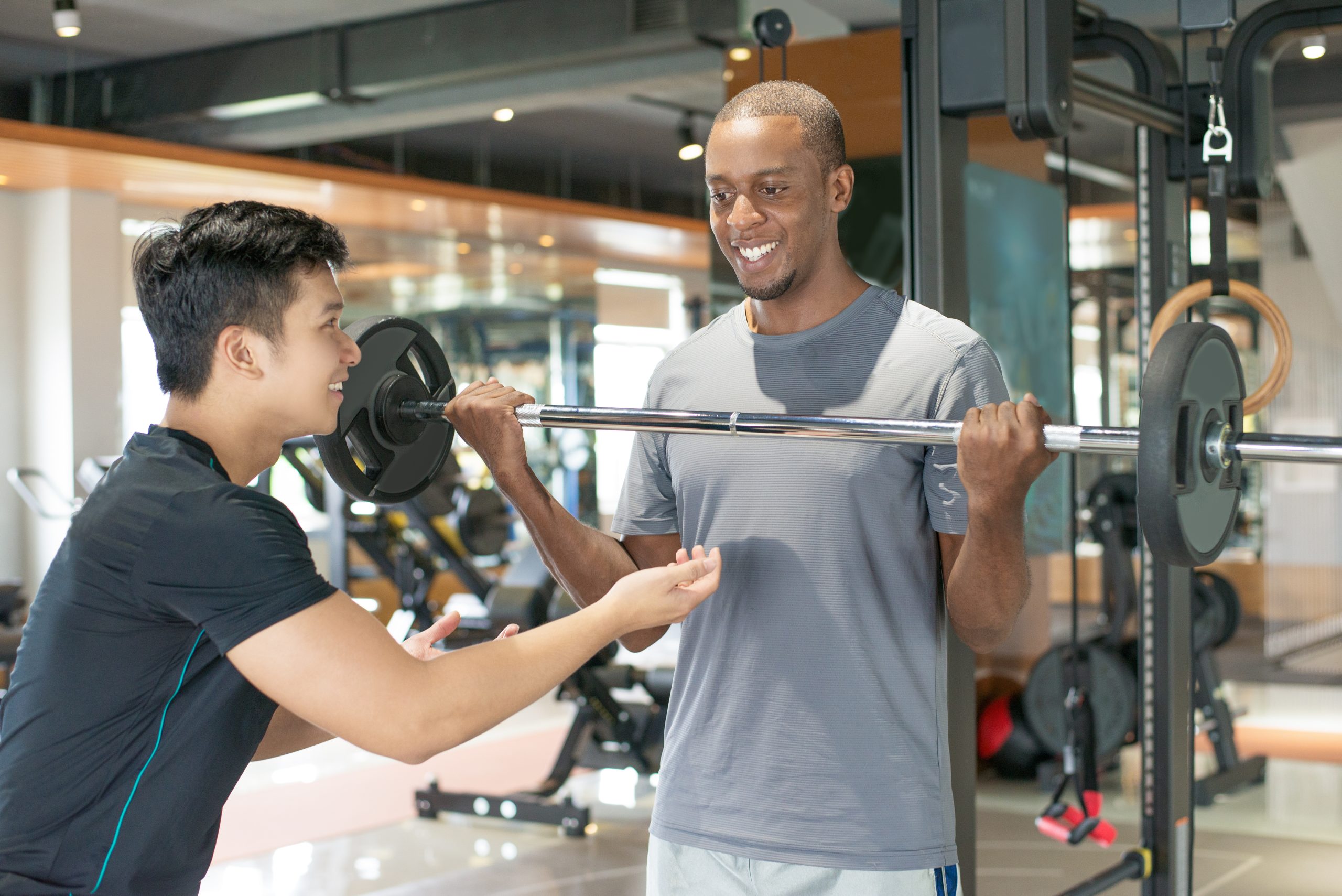 Smiling black man lifting barbell with personal trainer. Young guy training with gym equipment in background. Bodybuilding concept.